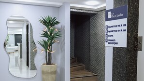 A reception area with a large mirror, a potted plant, and a sign indicating the location of the reception desk. - Hotel Place Jardin (Garanhuns)