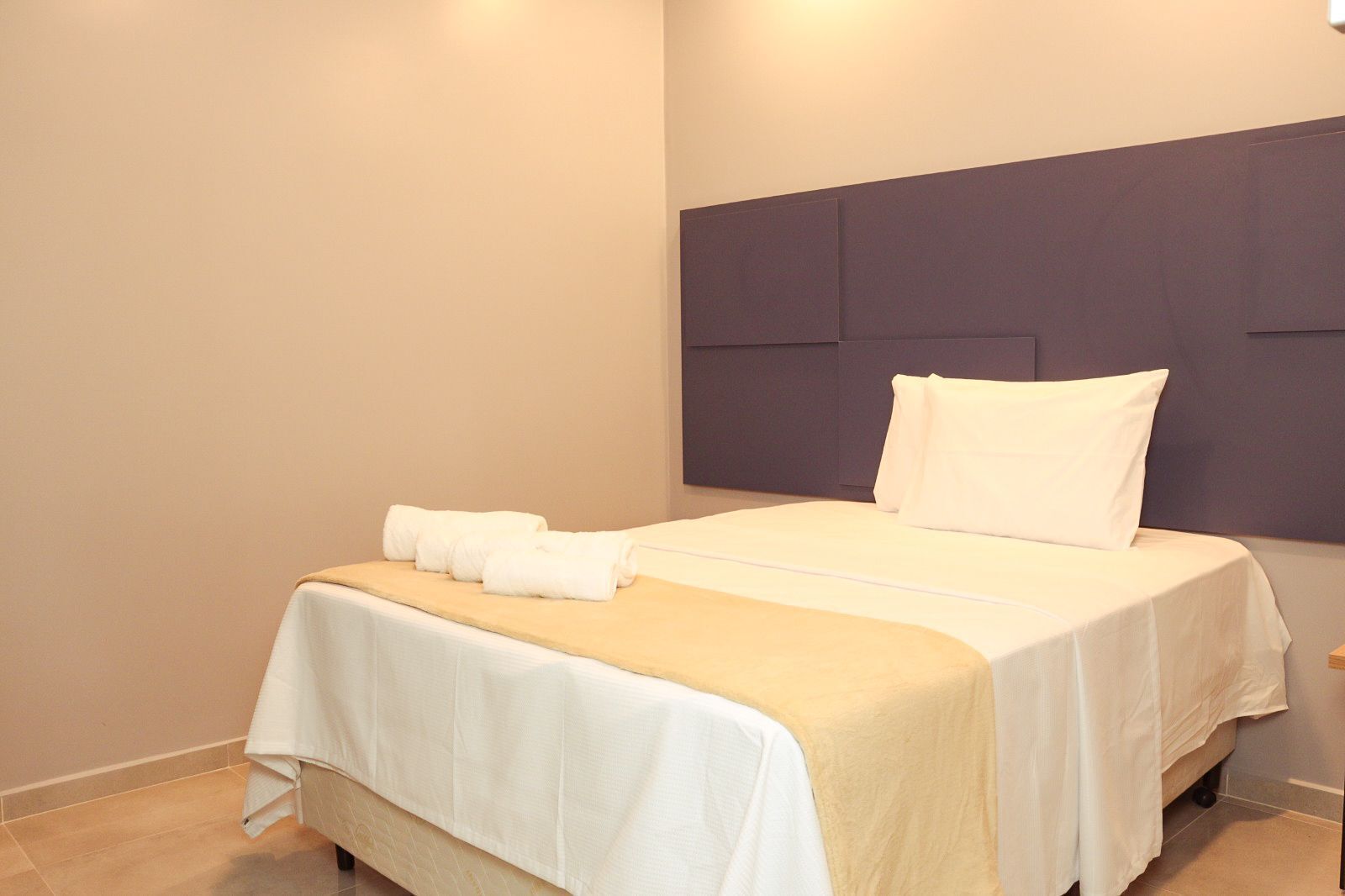 Una cama bien hecha con sábanas blancas, un plaid color beige y un cabecero con panel oscuro.