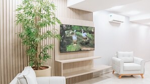 A modern living room with a flat-screen TV mounted on a wooden wall, a white sofa, a white armchair, and a large bamboo plant. - Hotel Place Jardin (Garanhuns)