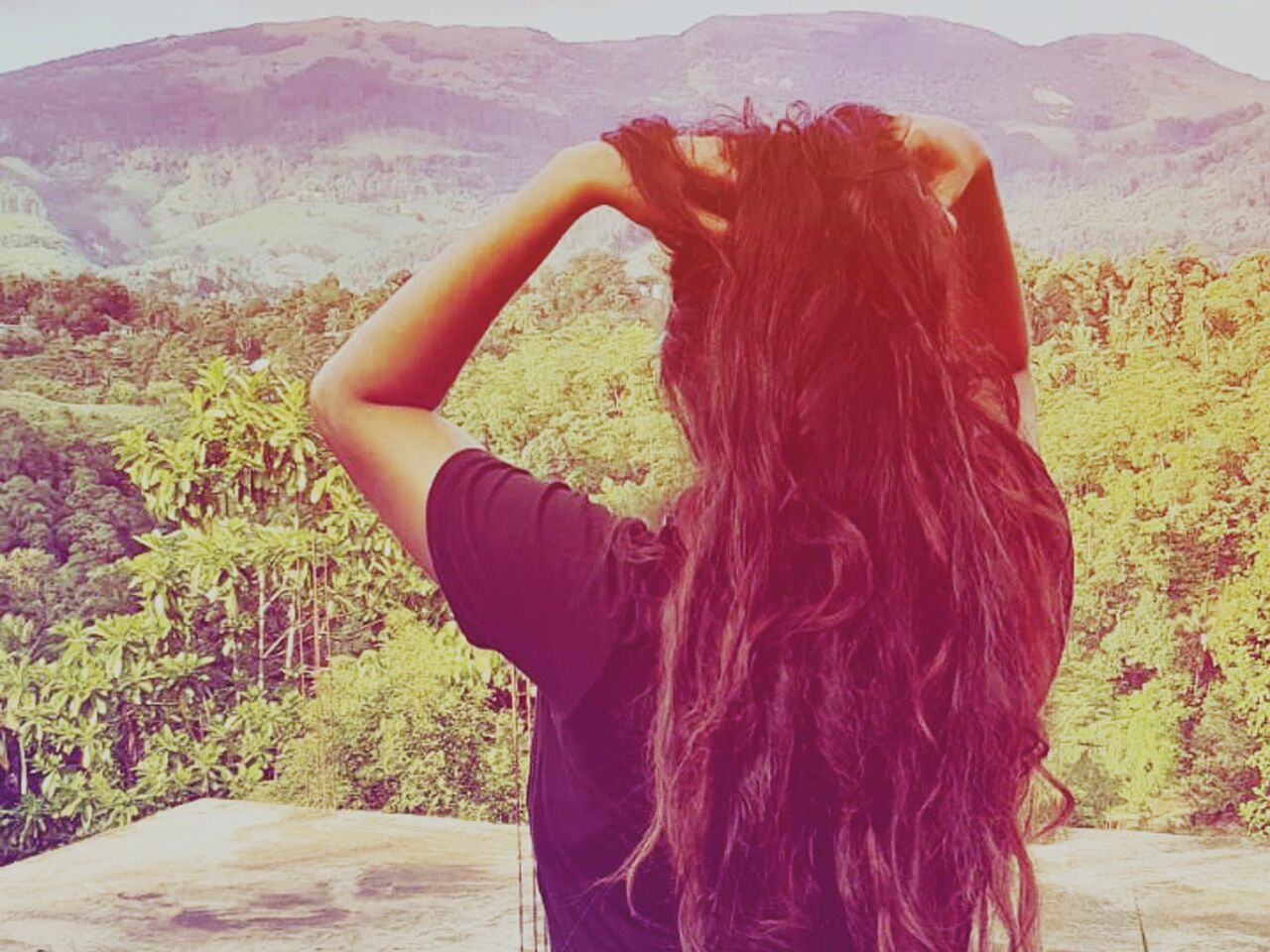 A person with long hair standing with their back to the camera, overlooking a mountainous landscape with greenery.