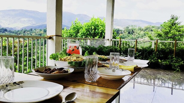 Mesa de jantar posta ao ar livre, com pratos e copos, rodeada por vegetação e montanhas ao fundo.