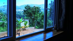 Quarto com uma janela ampla que oferece vista de um jardim exuberante e montanhas ao longe.