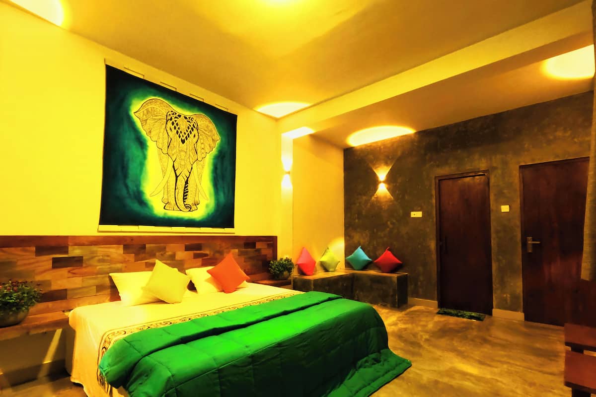 A hotel room with a bed, a large wall tapestry, a wooden headboard, and a small table with colorful cushions.
