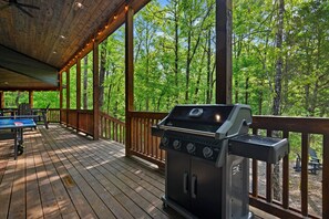 A covered deck with a barbecue grill, a ping pong table, and a view of a forest. - Family Fun Cabin Retreat w/ Arcade & Ping Pong (Broken Bow)