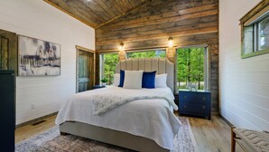 A bedroom with a wooden ceiling, a large bed with a white and beige quilt, two nightstands, and a view of trees through a window. - Family Fun Cabin Retreat w/ Arcade & Ping Pong (Broken Bow)