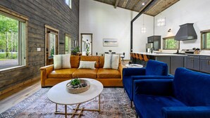 A modern living room with a brown leather sofa, a blue sofa, and a white coffee table. There is a kitchen area with a large window and a hanging light fixture. - Family Fun Cabin Retreat w/ Arcade & Ping Pong (Broken Bow)