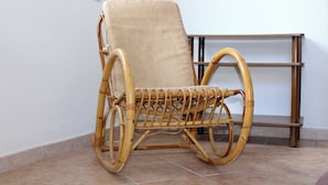 A wicker rocking chair with a beige cushion, positioned on a tiled floor next to a wooden shelf unit.