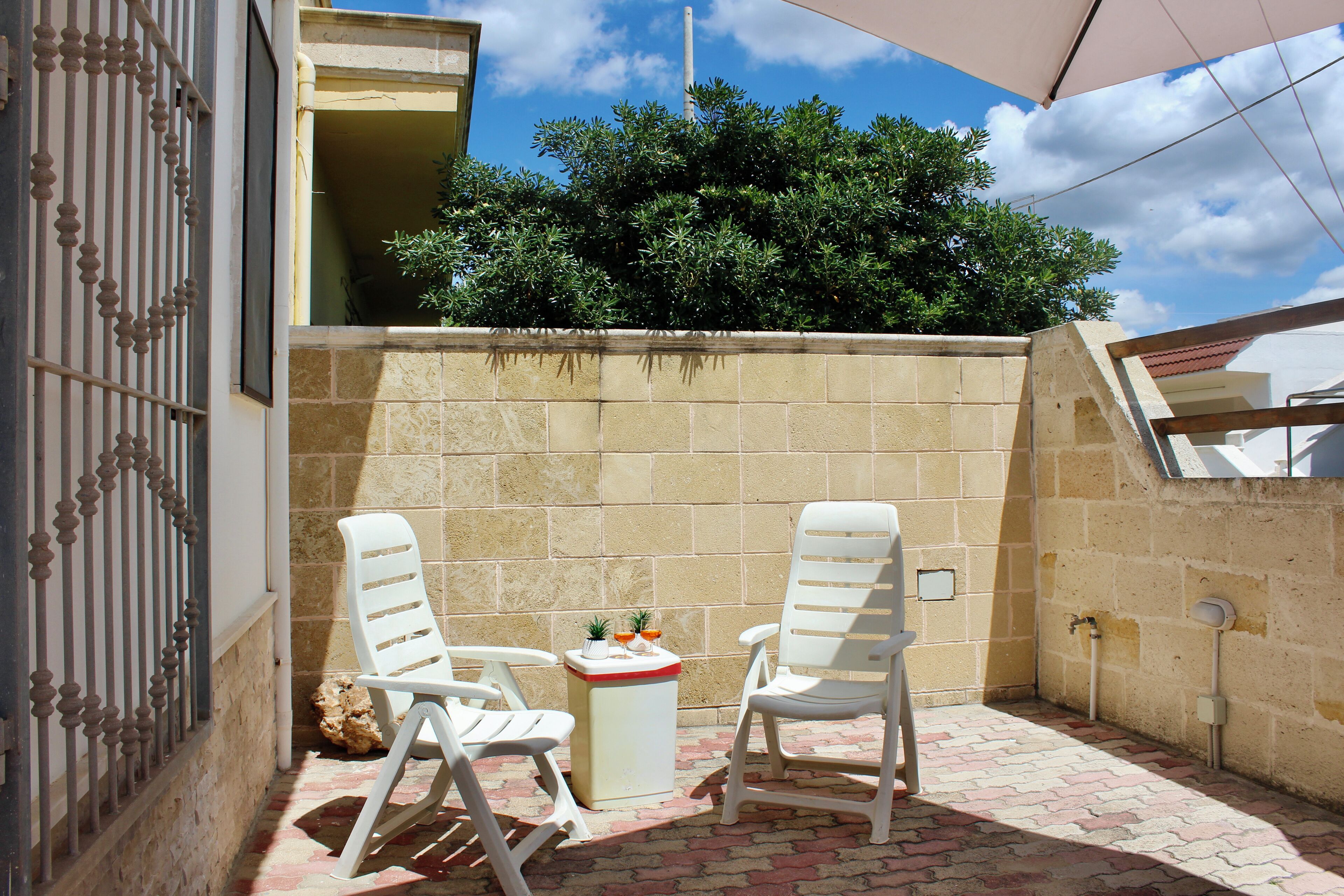 An outdoor patio with two white chairs, a small table with drinks, and a view of a building and trees.