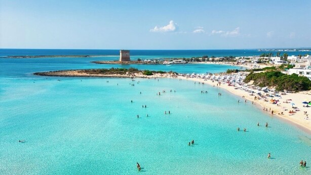 A beach with clear turquoise water, a sandy shore, and a tower in the distance.