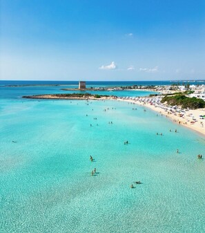 A beach with clear turquoise water, a sandy shore, and a tower in the distance.