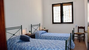 A bedroom with two single beds, a window with bars, a chair, and a nightstand with a lamp.