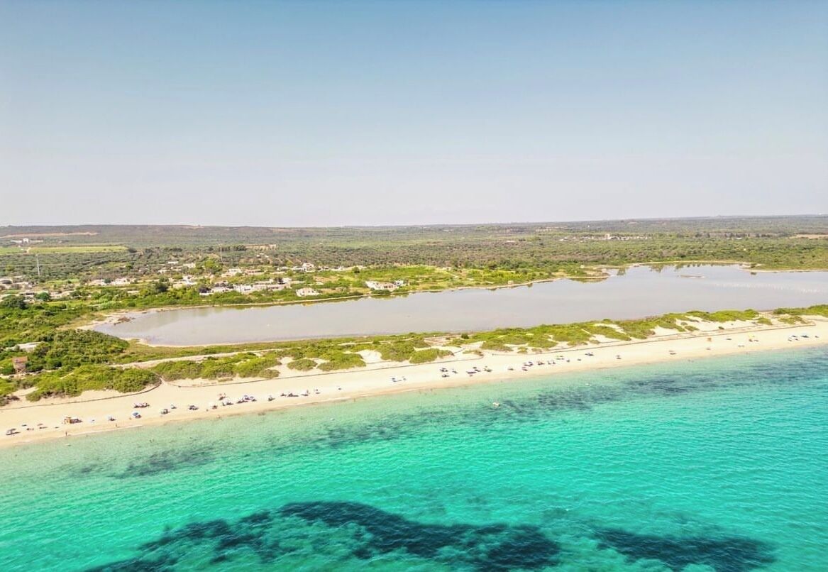 An aerial view of a coastal area with a beach, clear turquoise waters, and a small lake surrounded by greenery.