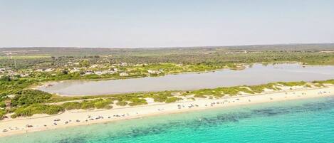 An aerial view of a coastal area with a beach, clear turquoise waters, and a small lake surrounded by greenery.