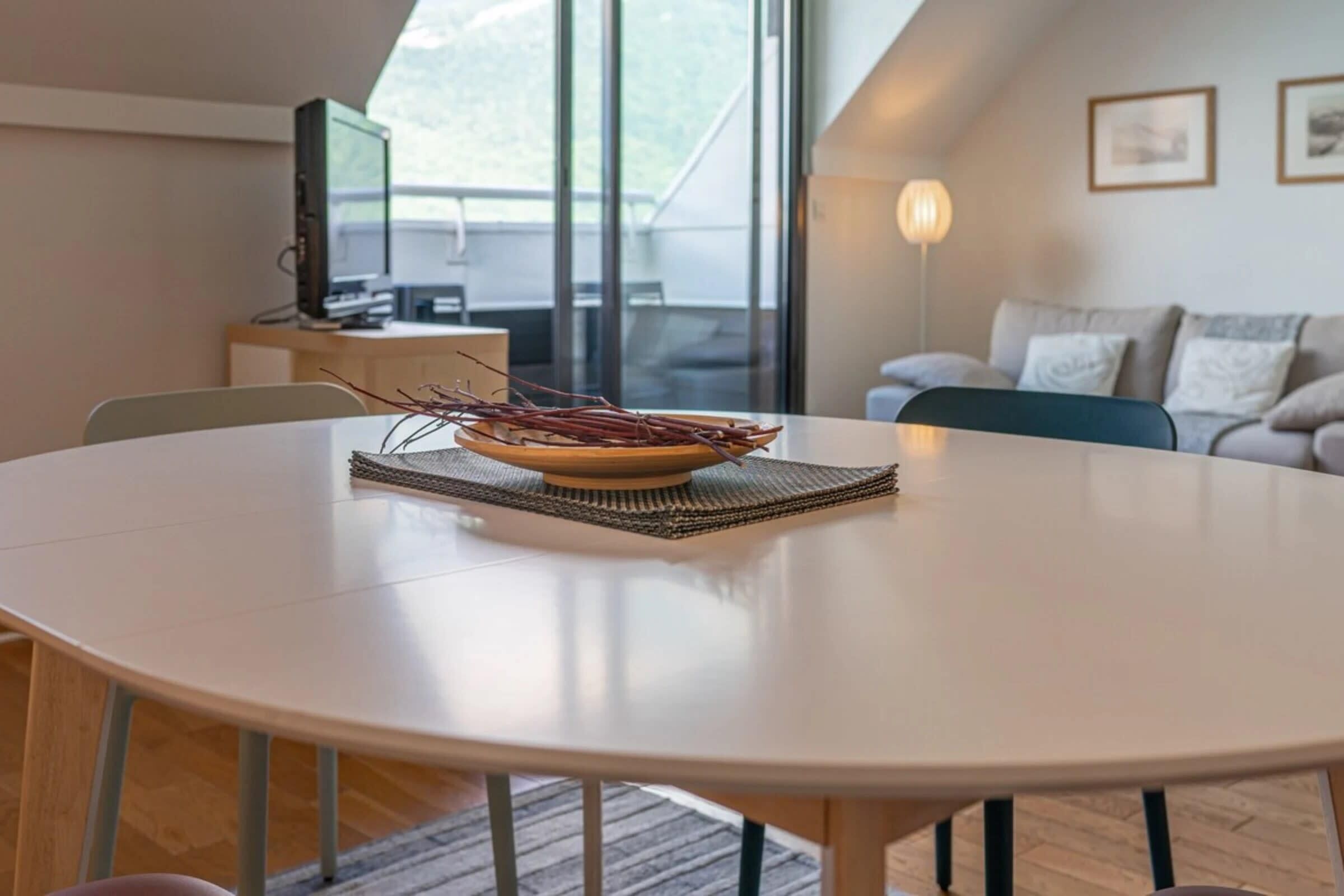 A round dining table with a bowl and branches on a woven placemat, a television on a desk, and a sofa in the background.