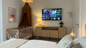 Et moderne soveværelse med et stort fladskærms-tv på væggen, en tremmeseng, et natbord med en lampe og en stor seng med et hvidt dynebetræk.