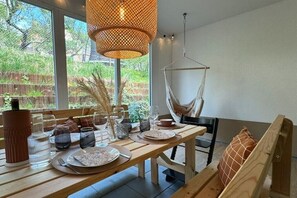 A dining area with a wooden table set for two, a hanging chair, and a large window with a view of greenery.