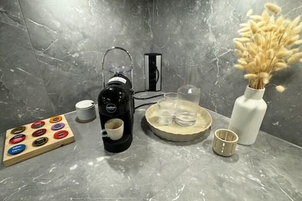 A bathroom with a coffee maker, a tea kettle, a wooden board with tea bags, a vase with dried flowers, and a tray with glasses.