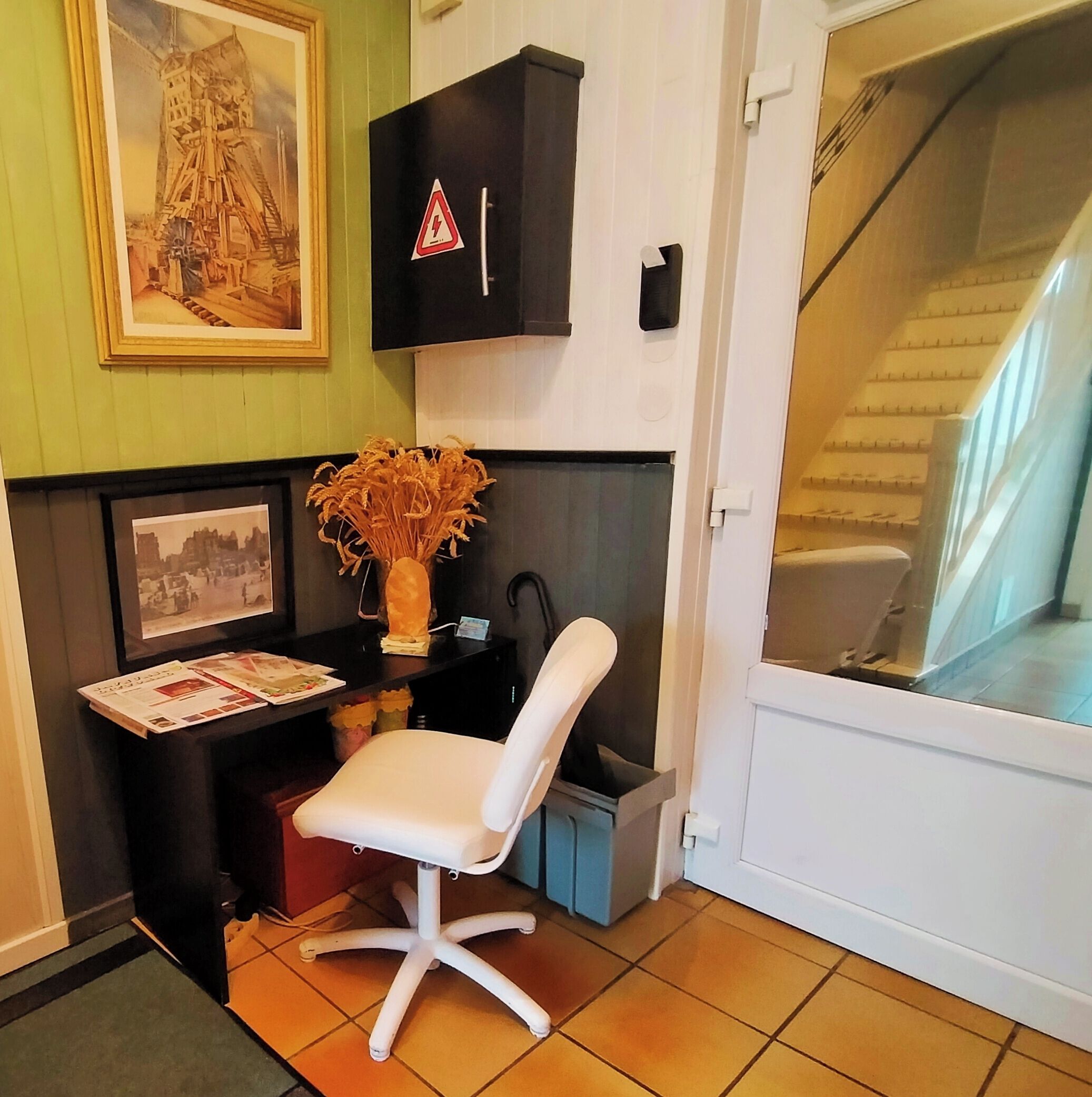 A room with a desk, chair, framed pictures, a warning sign, and a staircase.