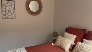 Un dormitori amb un edredó vermell, una manta blanca amb textura i coixins decoratius. Un mirall rodó penja de la paret, i es pot veure una obra d'art emmarcada.