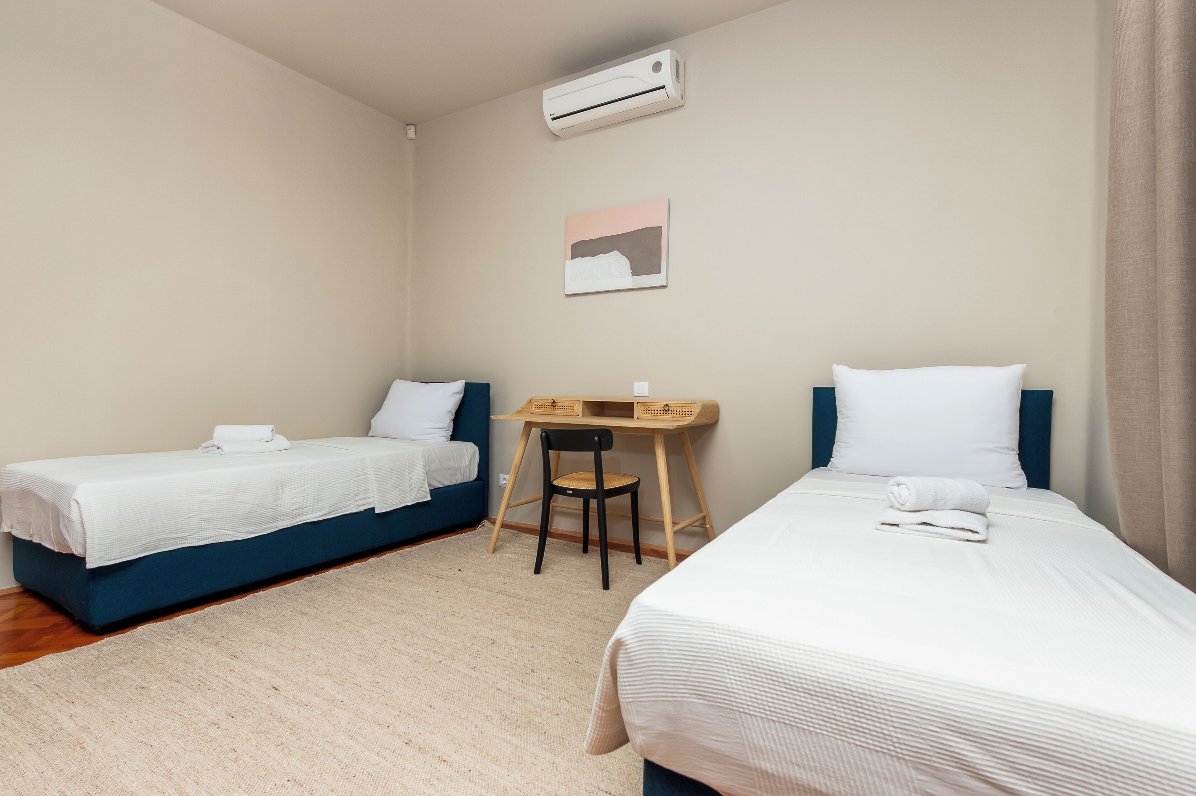 A hotel room with two single beds, a desk, a chair, an air conditioner, and a painting on the wall.