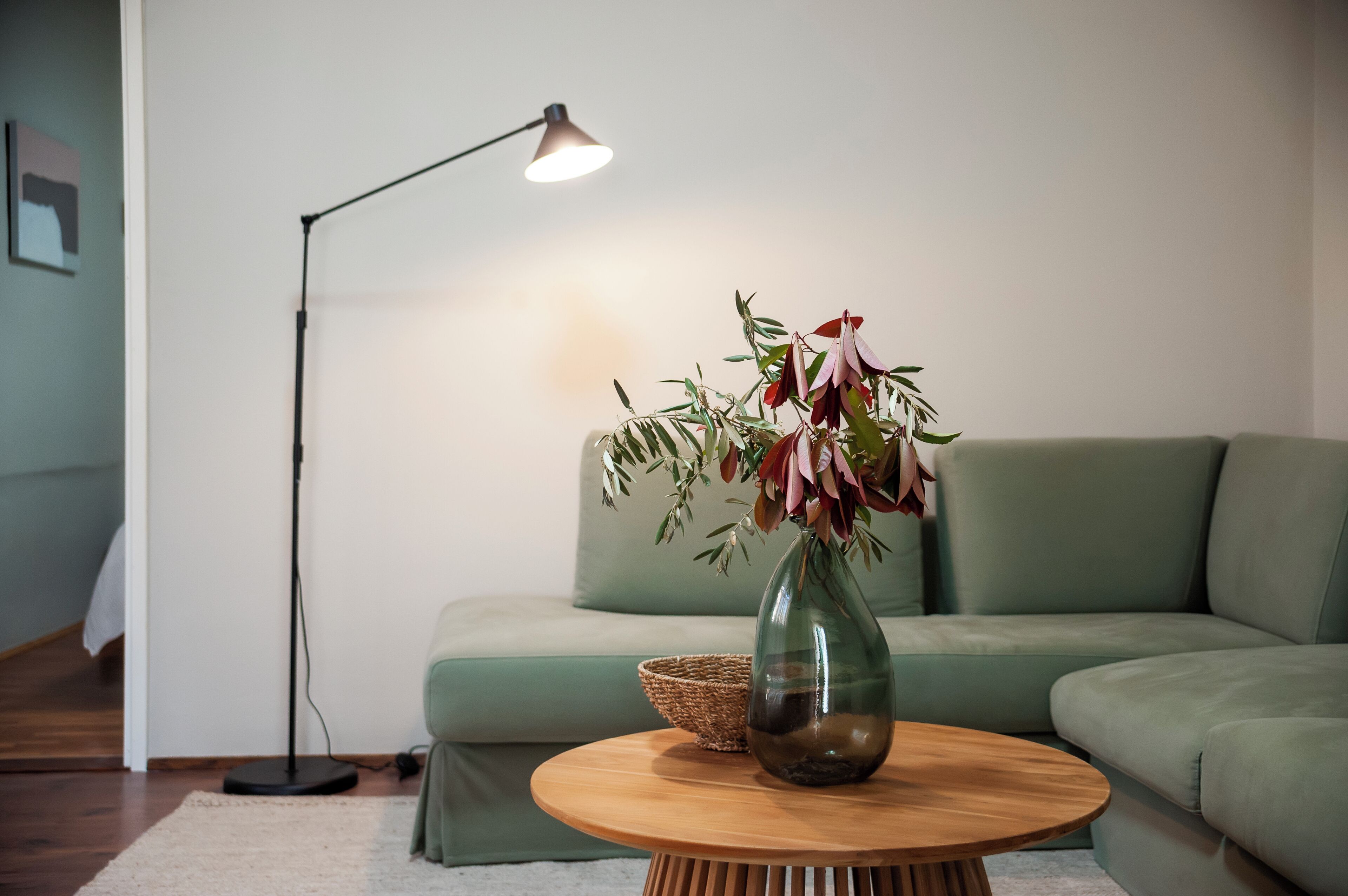 Una sala de estar con un sofá verde, una mesa de centro de madera con un florero, una lámpara de pie y una cesta tejida.