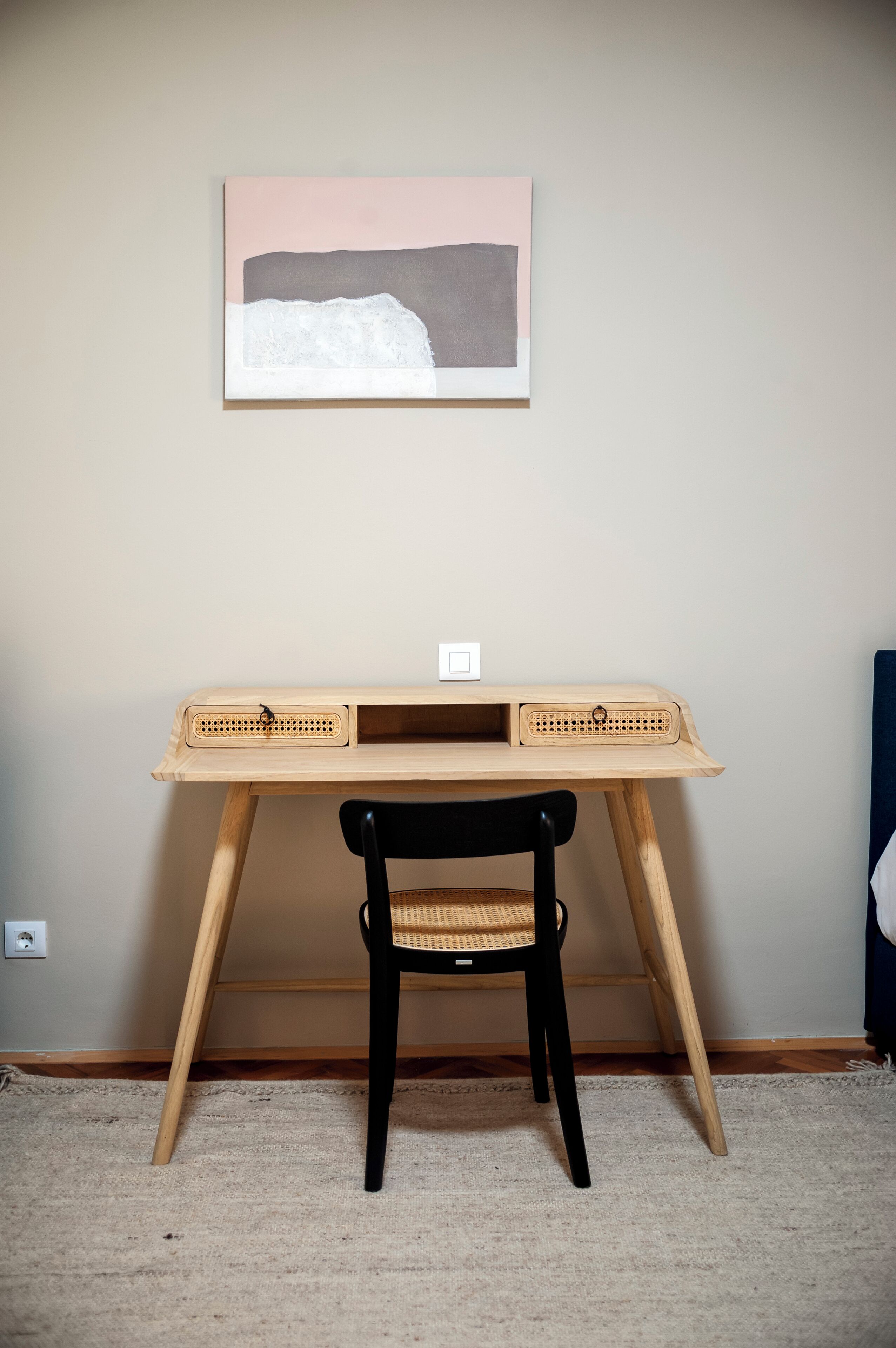 Un escritorio de madera con una silla tejida, un cuadro en la pared y un enchufe.