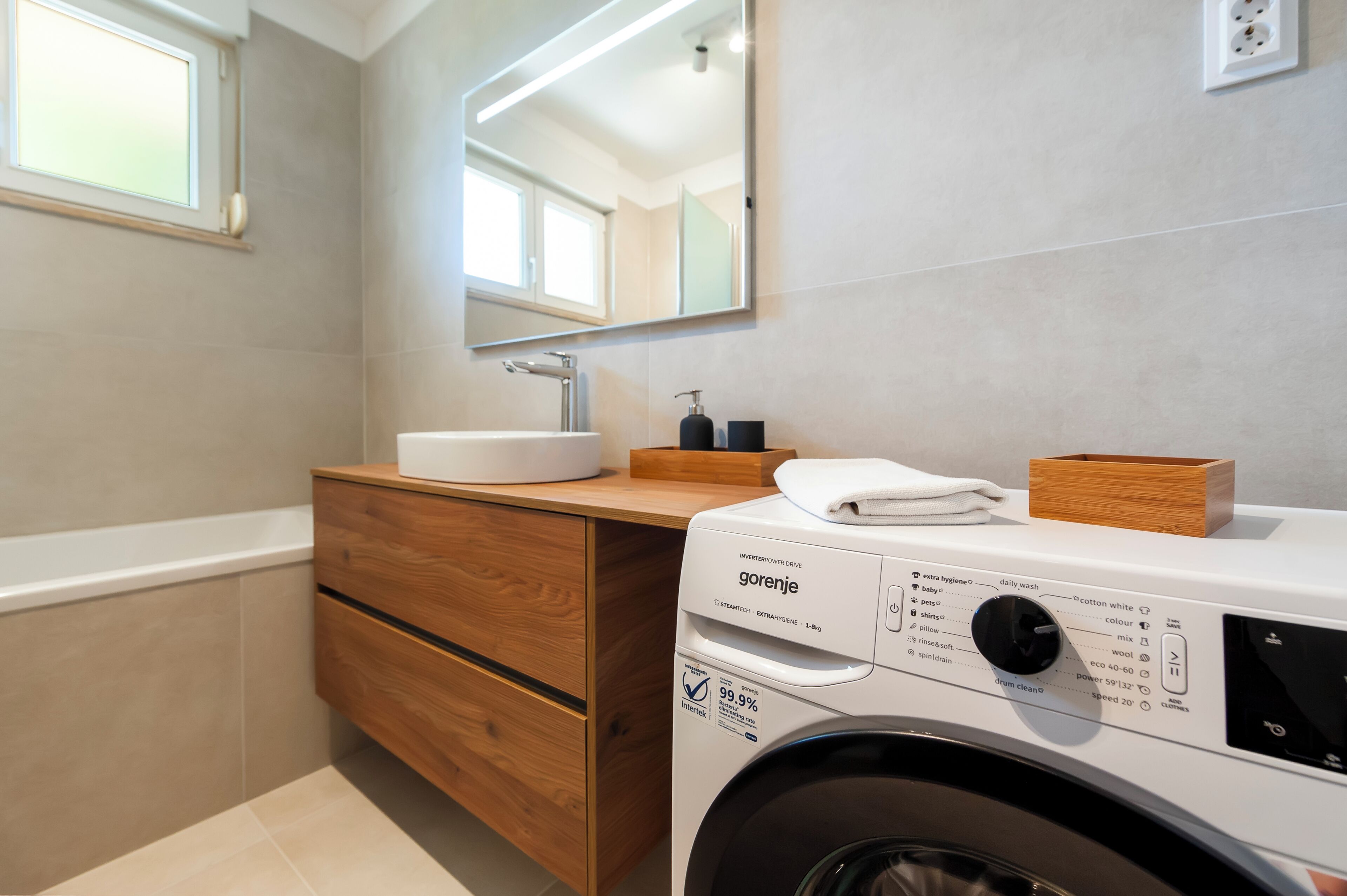 Een moderne badkamer met een houten wastafelmeubel, een witte wastafel en een voorlader wasmachine.