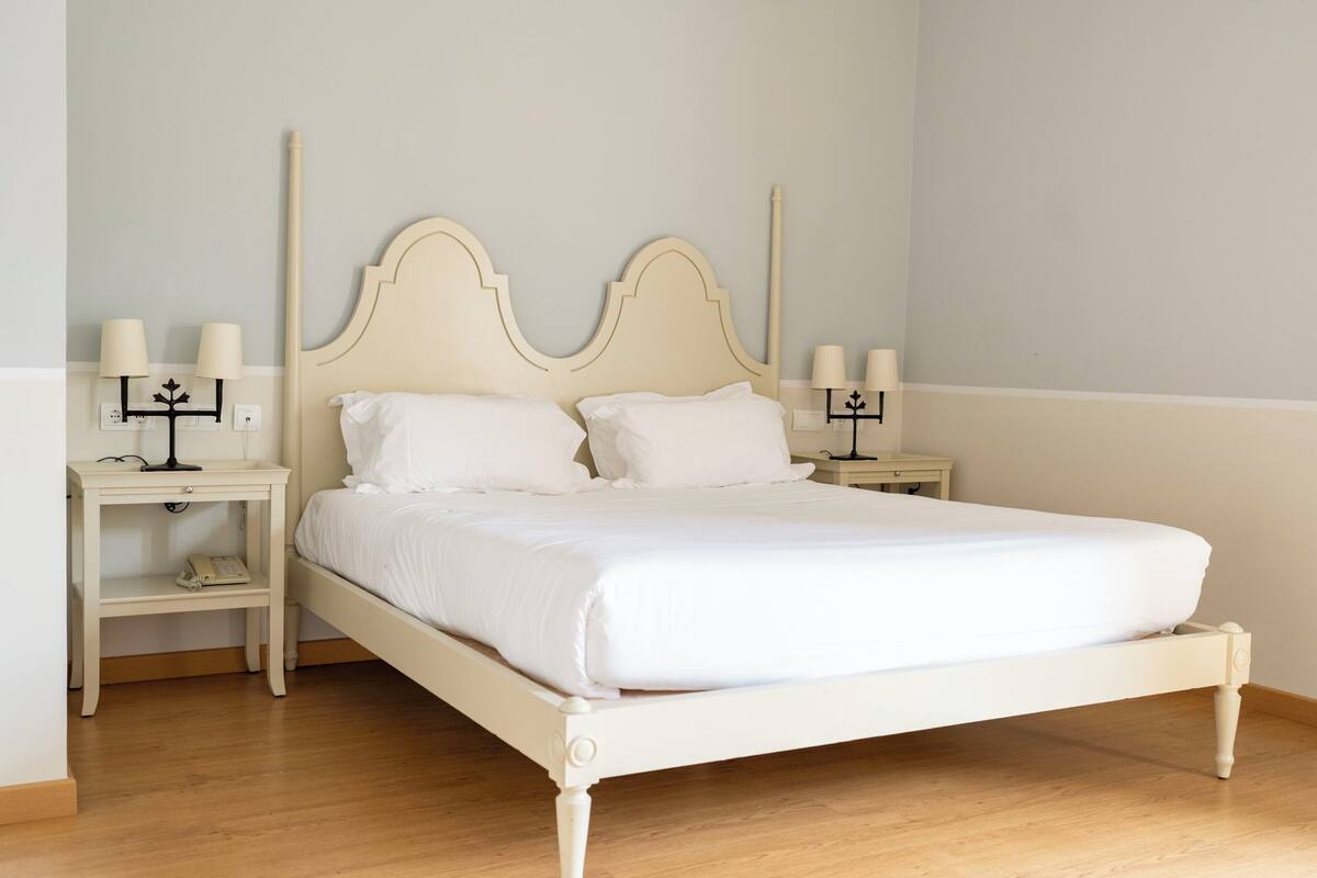 A white bed with a wooden frame in a room with light-colored walls and wooden flooring.
