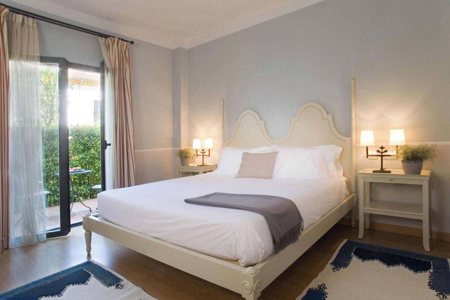 Una cama bien hecha con sábanas blancas y un plaid gris, dos mesitas de noche con lámparas, una planta en maceta y vistas a un balcón con cortinas.