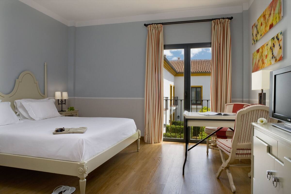 A hotel room with a bed, a desk, a chair, a TV, and a balcony with curtains.