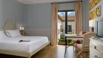 Habitación de hotel con cama, escritorio, silla, televisor y balcón con cortinas.