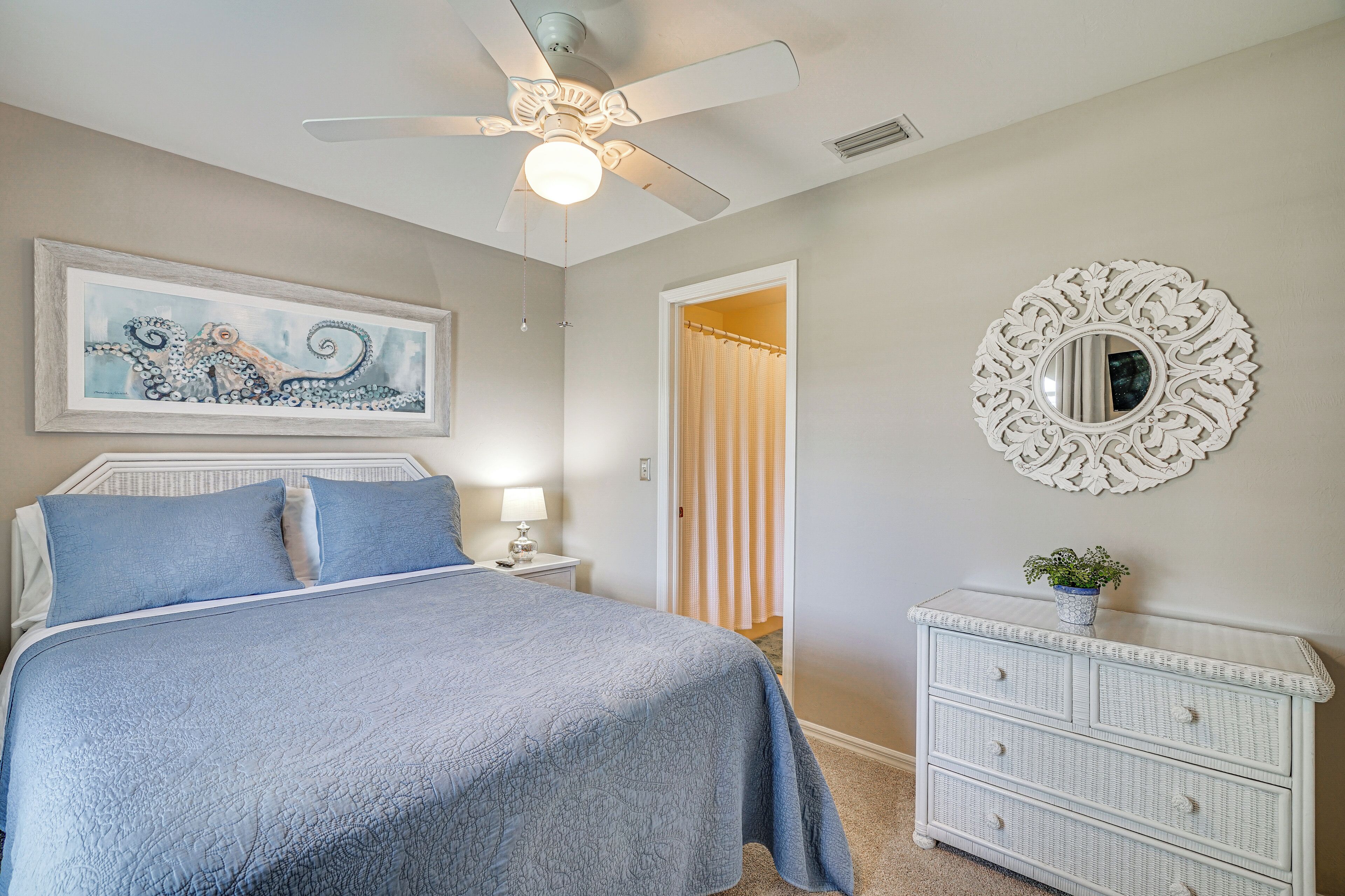 A bedroom with a bed, a ceiling fan, a wall-mounted mirror, and a painting.
