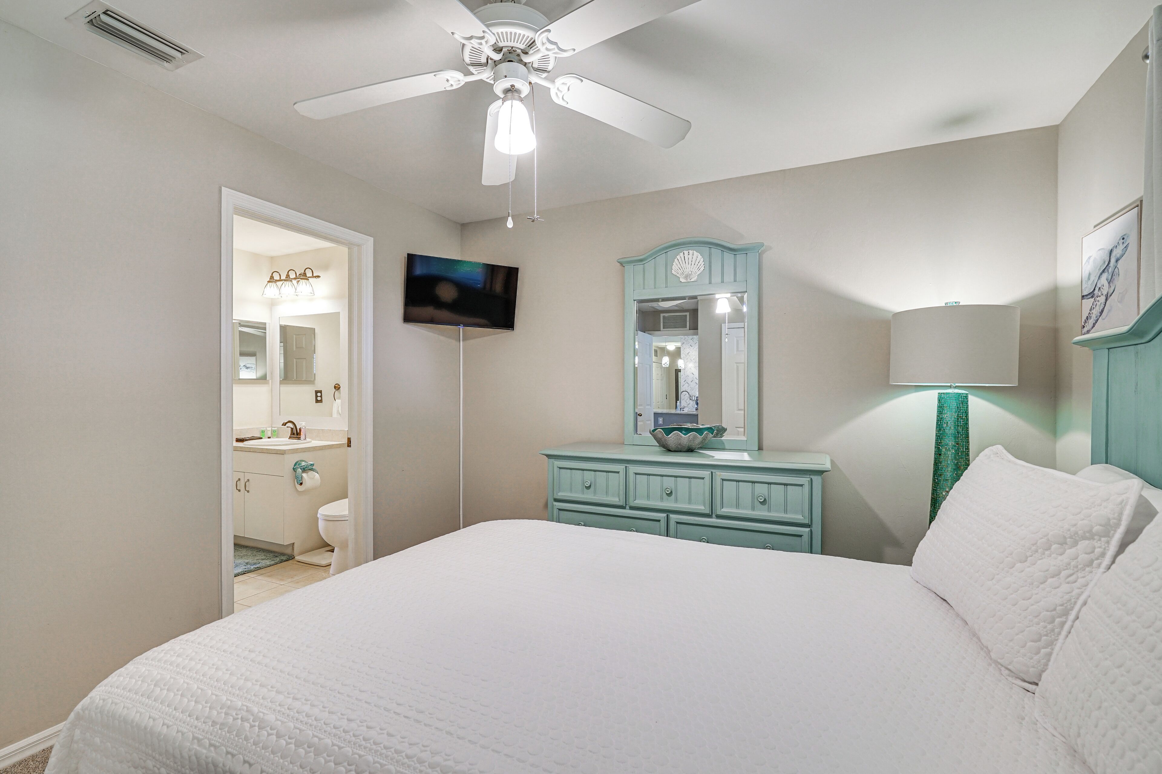 Une chambre à coucher comprenant un lit, un ventilateur de plafond, une télévision, une coiffeuse avec un miroir et une table de chevet avec une lampe.