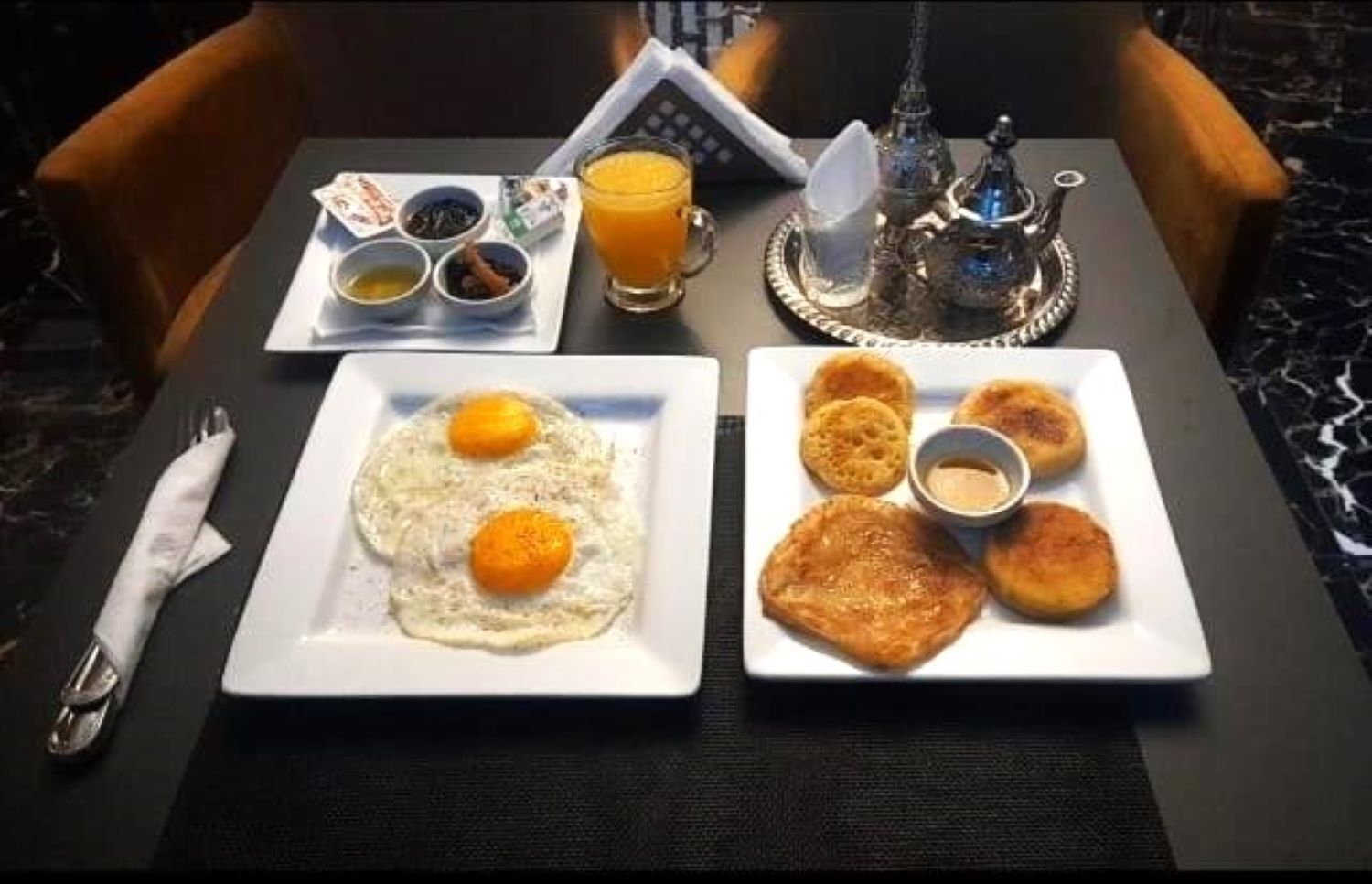 Una escena de desayuno con dos platos de comida, un vaso de jugo de naranja y una tetera sobre una mesa.
