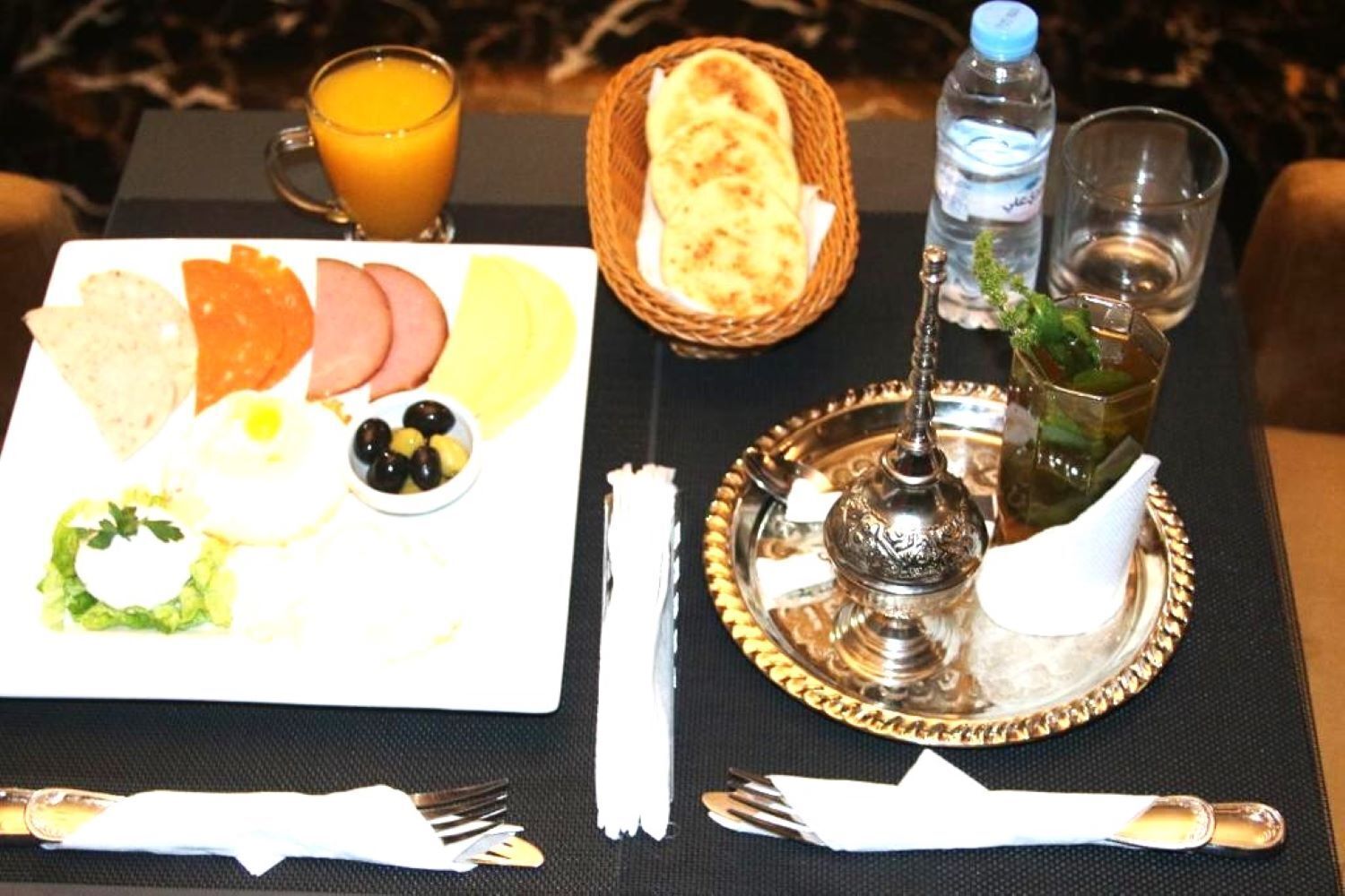 A breakfast setting with a plate of food, a glass of orange juice, a bottle of water, a basket of bread, and a hookah on a table.