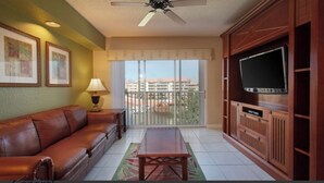 A living room with a leather sofa, a coffee table, a TV on a wooden cabinet, a ceiling fan, and a balcony with a view of buildings.