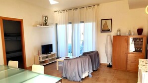 Une pièce comprenant une table à manger en verre, un téléviseur posé sur un meuble, un lit recouvert d’une couverture grise, une armoire en bois et une fenêtre avec des rideaux.