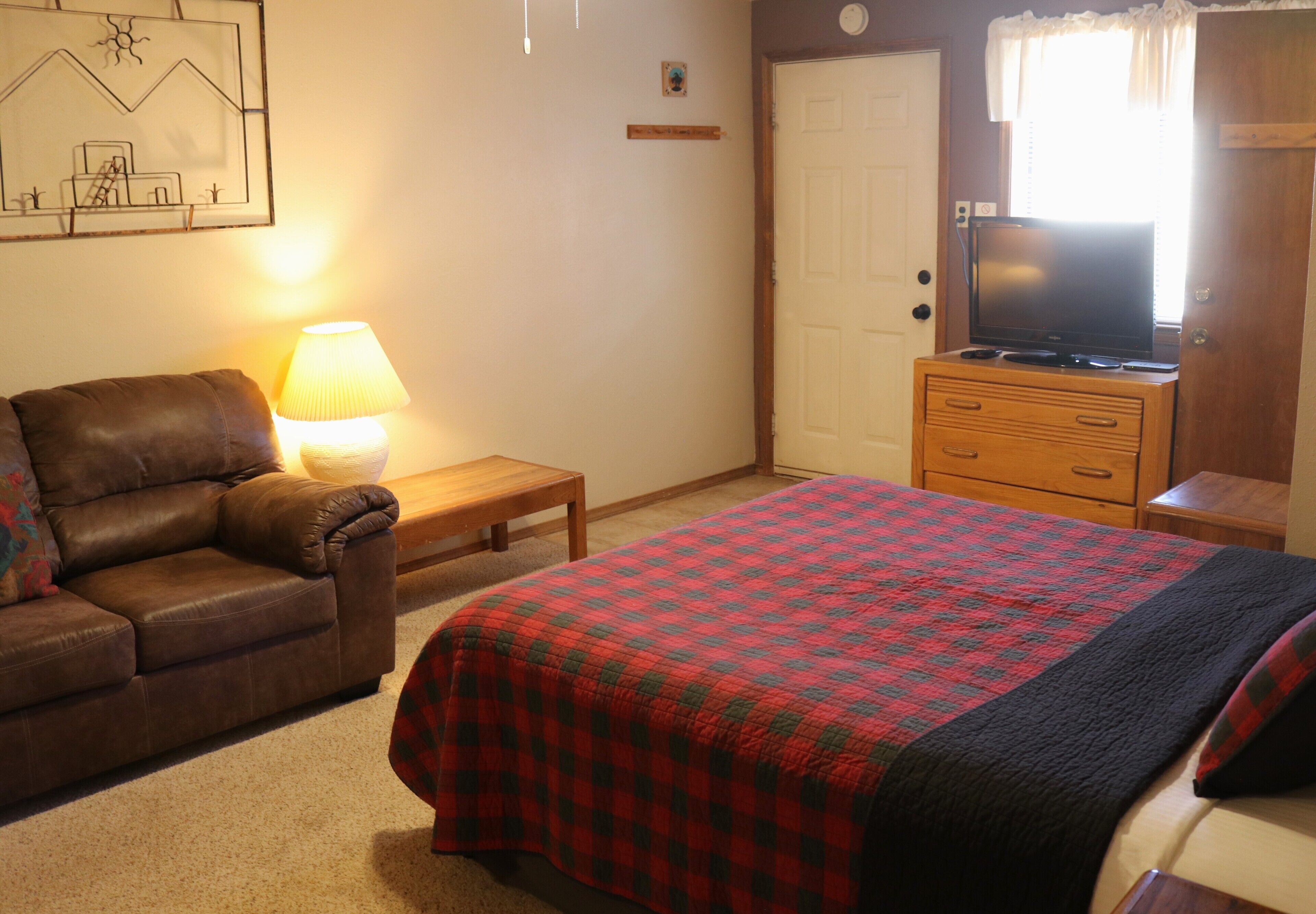 A bedroom with a bed, a wooden dresser, a television on top of the dresser, a lamp on a bedside table, and a brown leather couch.