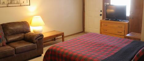 A bedroom with a bed, a wooden dresser, a television on top of the dresser, a lamp on a bedside table, and a brown leather couch.
