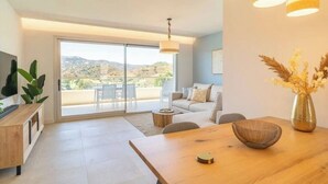 Ein modernes Wohnzimmer mit einem Holzesstisch, einer Couch und einem Fernseher auf einer Holzkonsole. Eine große Schiebeglasdoor führt zu einer Terrassenanlage im Freien.