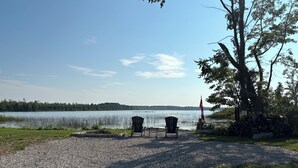 A gravel area with two Adirondack chairs, a flagpole, and a stone structure by a lake. - Welcome to the Lake House (Wiarton)