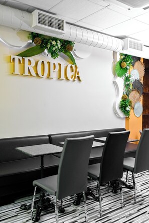 Ein Restaurantinneres mit einer Wandtafel mit dem Schriftzug „TROPICA“, einer Speiseauslage mit einer dekorativen Pflanzengestaltung und einem Sitzbereich mit Tischen und Stühlen.