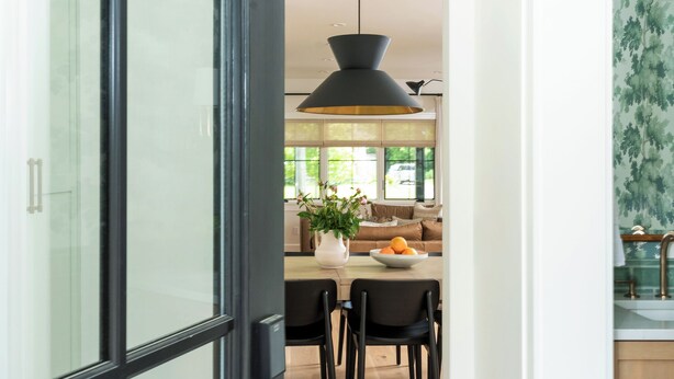 A modern kitchen with a dining area, featuring a black-framed glass door, a pendant light, and a view of the dining area through the door.