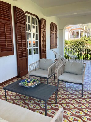 Een balkon met twee rieten stoelen, een tafel met een decoratief bord en vloertegels met een patroon.