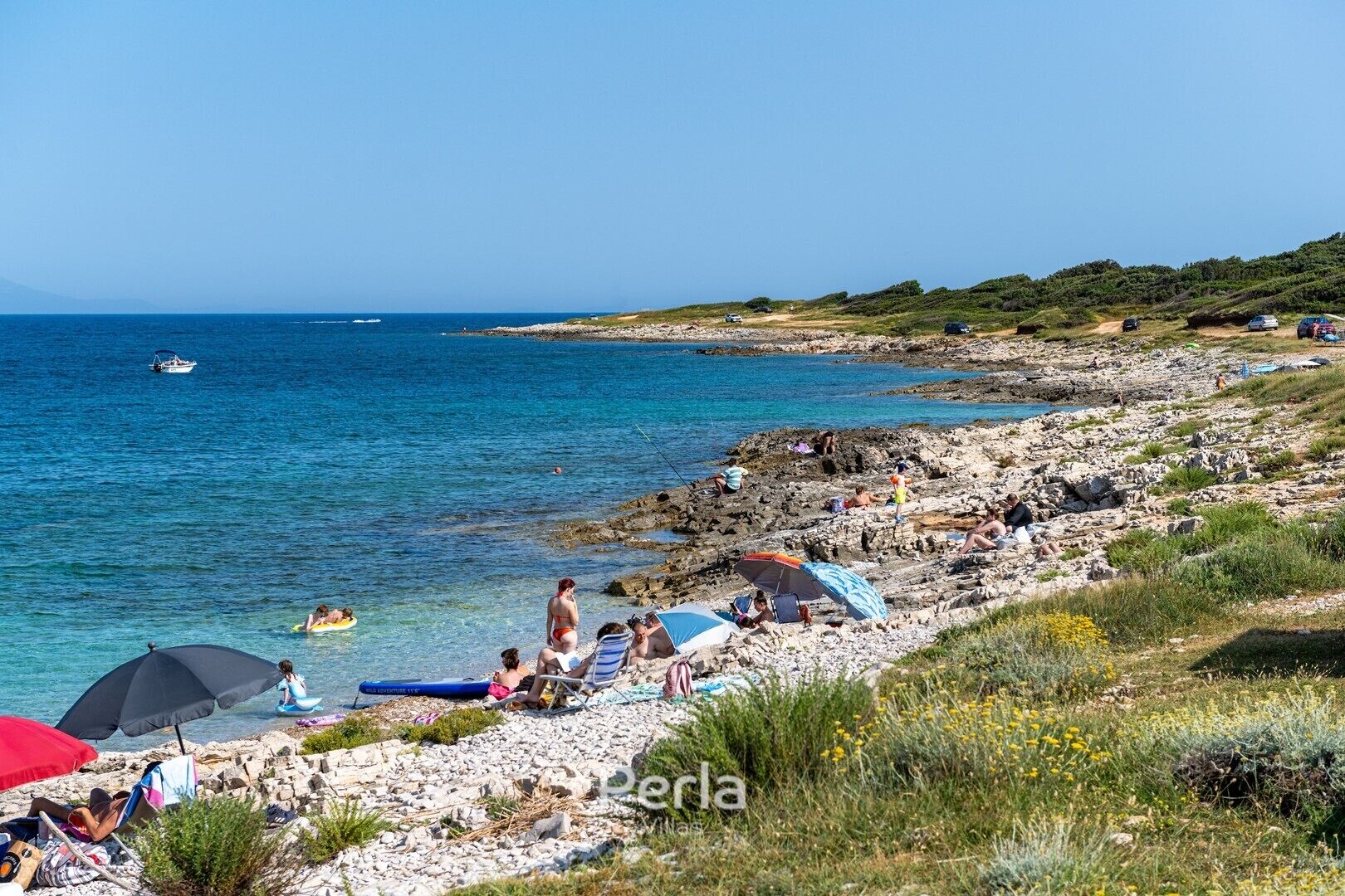A rocky beach with clear blue water, people sunbathing, and a boat in the distance.