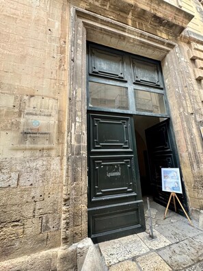 A building entrance with a large black door, a sign indicating '4 Balconies Paladini Rooms,' and a small easel with a poster on the ground.