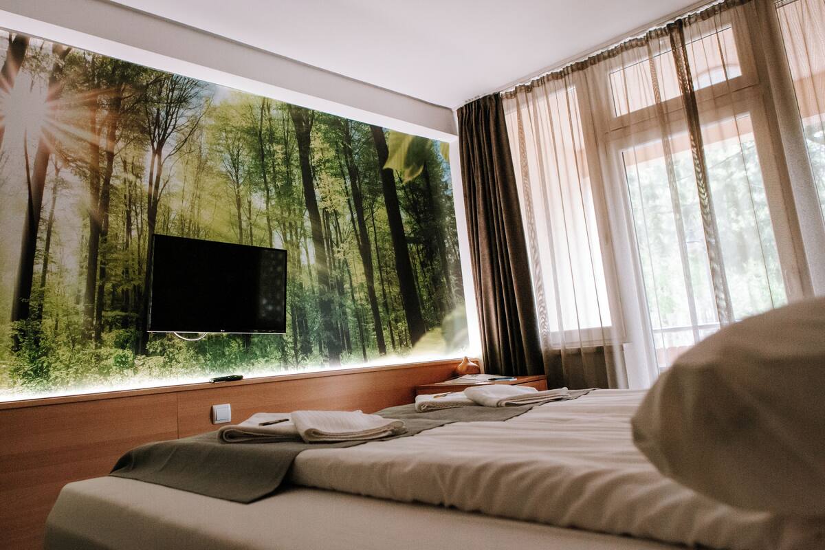 Une chambre d’hôtel avec un grand décor mural représentant une forêt, un téléviseur à écran plat, un lit avec des serviettes soigneusement pliées et une fenêtre ornée de rideaux légers.