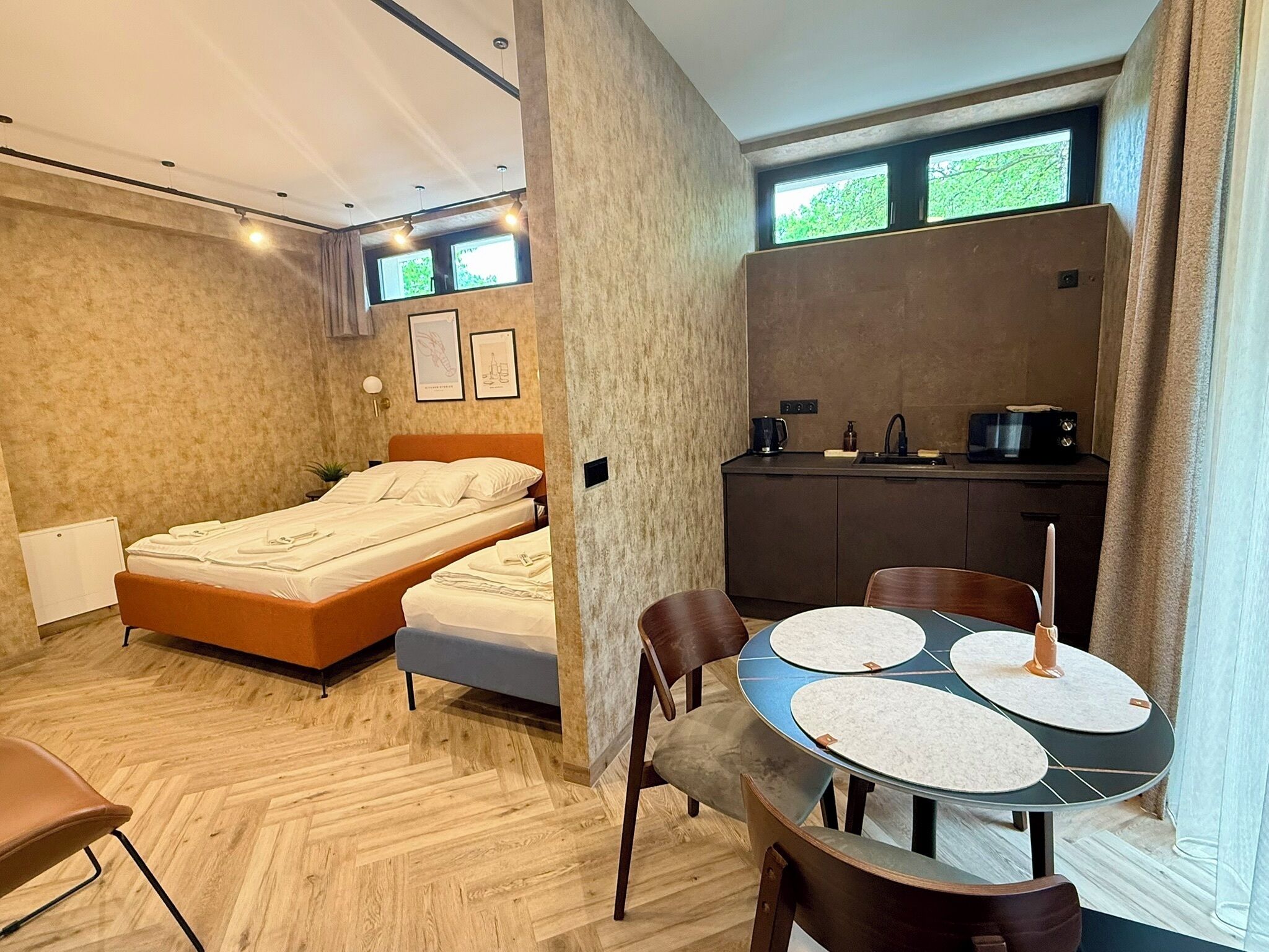 A compact room with a bed, a kitchenette, a dining table with chairs, and a window with a view of greenery.
