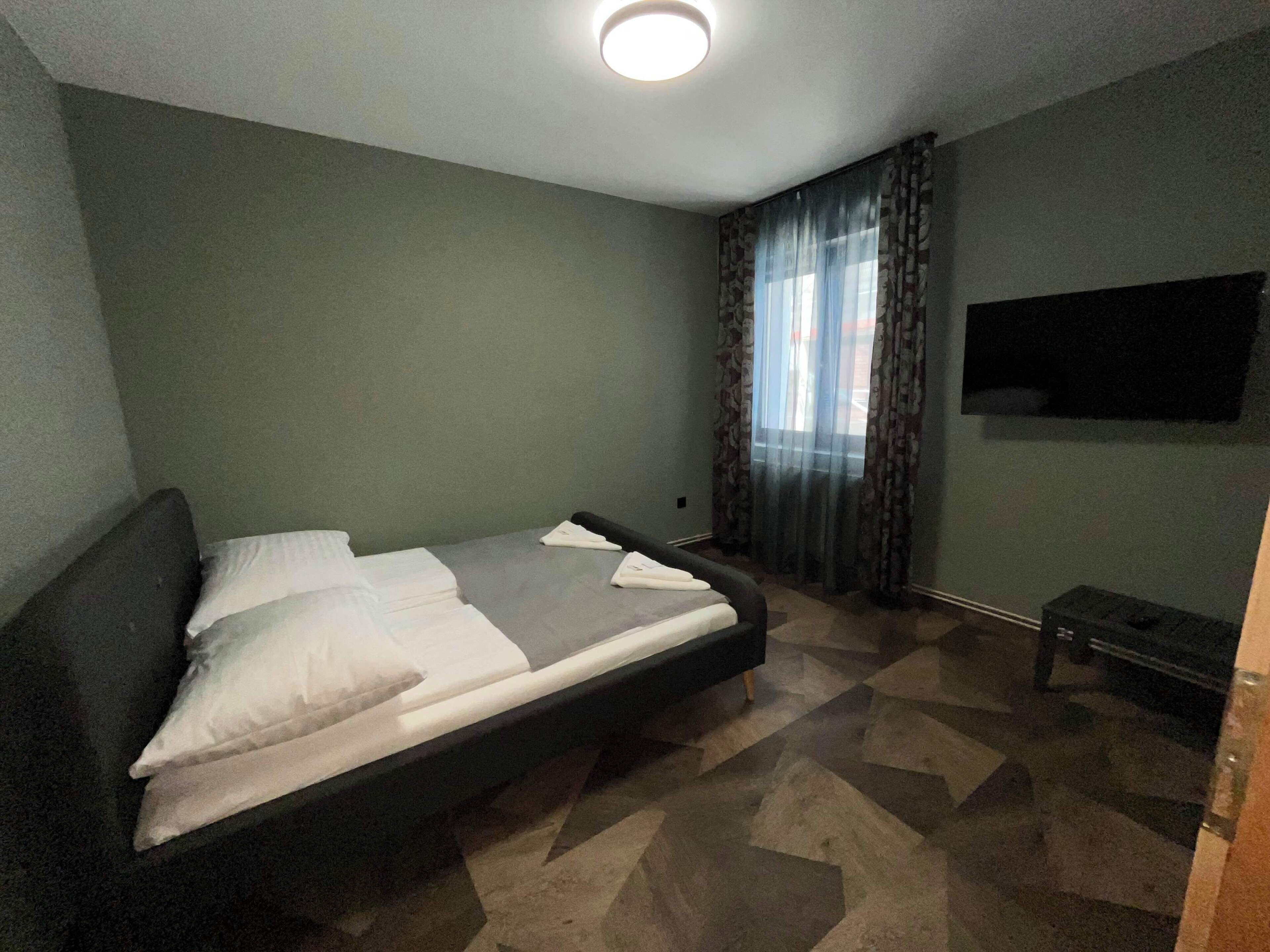Una cama individual con almohadas, una mesita de noche, un televisor montado en la pared y una ventana con cortinas.