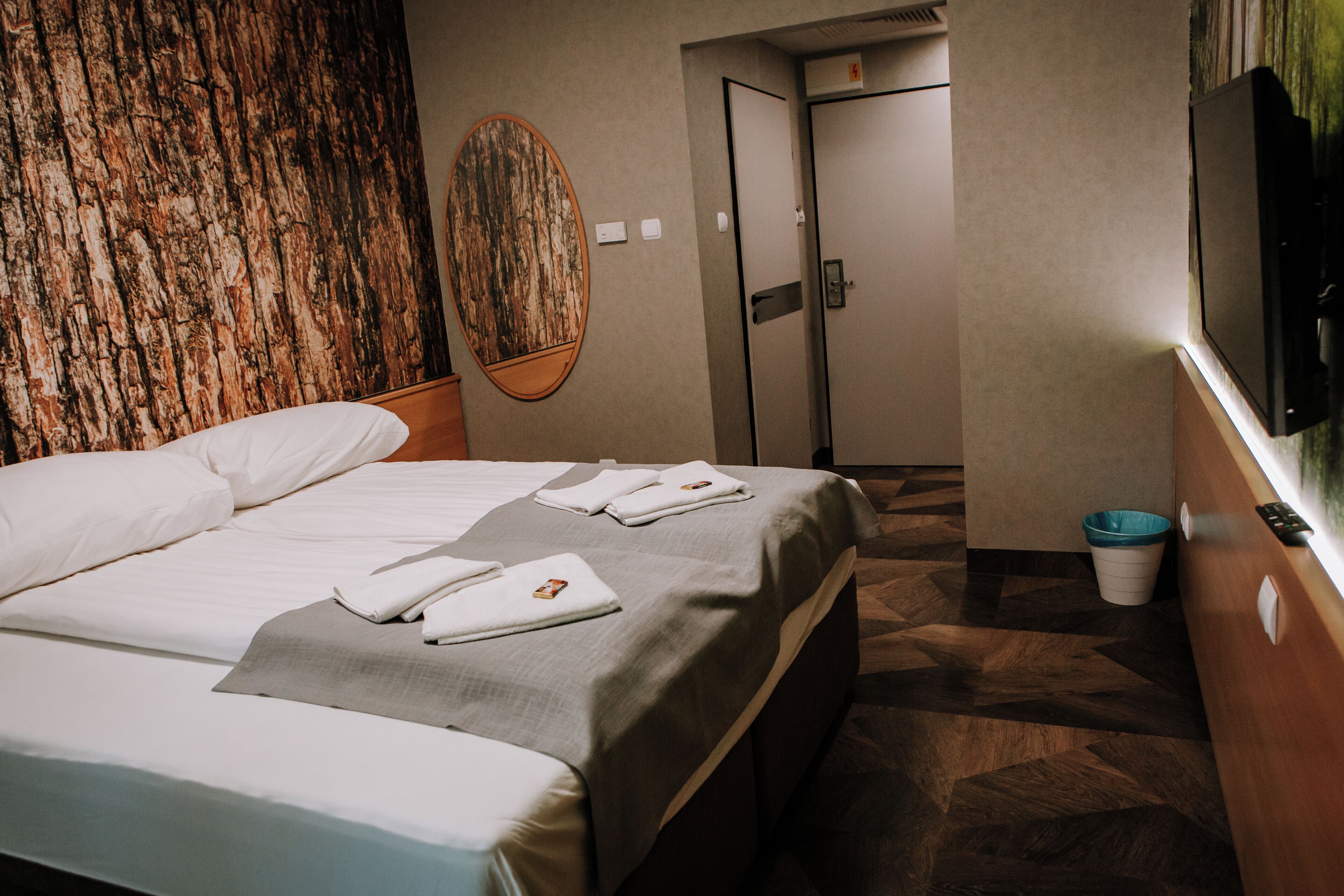 Habitación de hotel con cama, televisor montado en la pared, una puerta y un cubo de basura.